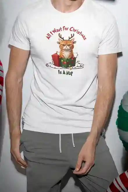 A man wearing a white t-shirt with an image of an angry cat with reindeer antlers sitting in a Christmas present. The text reads, "All I want for Christmas is a nap." He is standing with his hands in the pockets of his grey pants.  | Satire Clothes