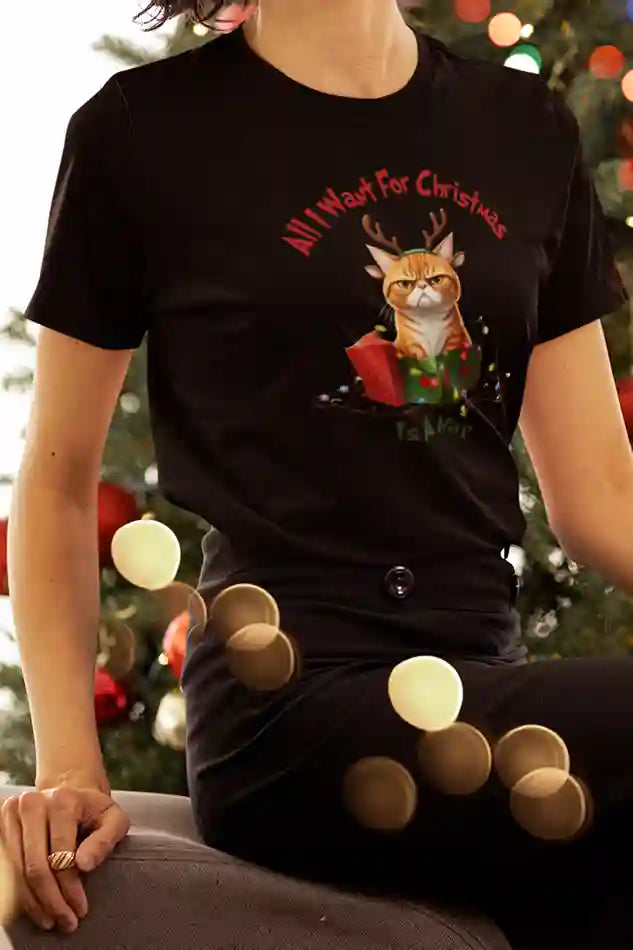 A woman wearing a black t-shirt with an image of an angry cat with reindeer antlers sitting in a Christmas present. The text reads, "All I want for Christmas is a nap." She is sitting in front of a decorated Christmas tree with colorful lights and ornaments.  | Satire Clothes