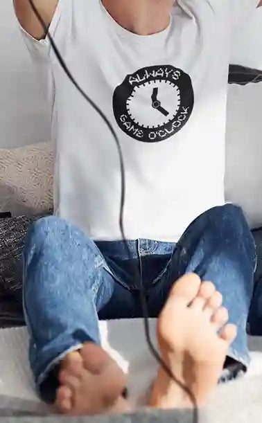 Man wearing a white t-shirt with a black circular graphic that says 'ALWAYS GAME O'CLOCK,' sitting with legs extended and bare feet visible, holding a game controller. | Satire Clothes