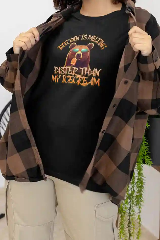 A woman wearing a black t-shirt with a bear holding an ice cream and the text "BITCOIN IS MELTING FASTER THAN MY ICECREAM" in orange letters. She has an open brown and black plaid flannel shirt over the t-shirt and is wearing light-colored cargo pants. | Satire Clothes