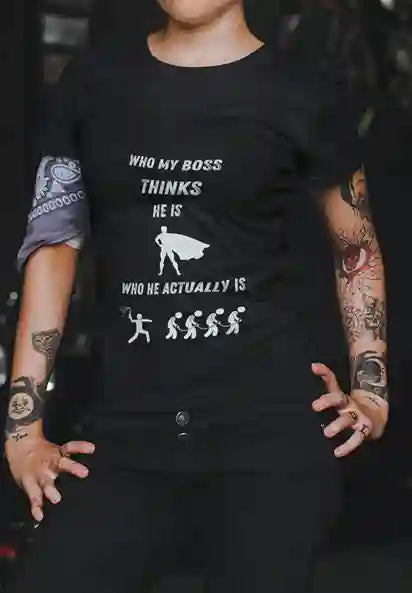 Woman wearing black satirical t-shirt with 'WHO MY BOSS THINKS HE IS' above a super hero silhouette, and 'WHO HE ACTUALLY IS' above a person whipping others | Satire Clothes