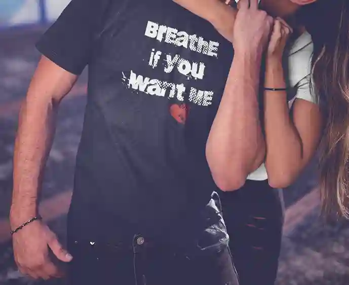 A man wearing a black t-shirt with white text that reads "BREATHE if YOU Want ME" and a red broken heart at the bottom. A woman has her arm around him. | Satire Clothes