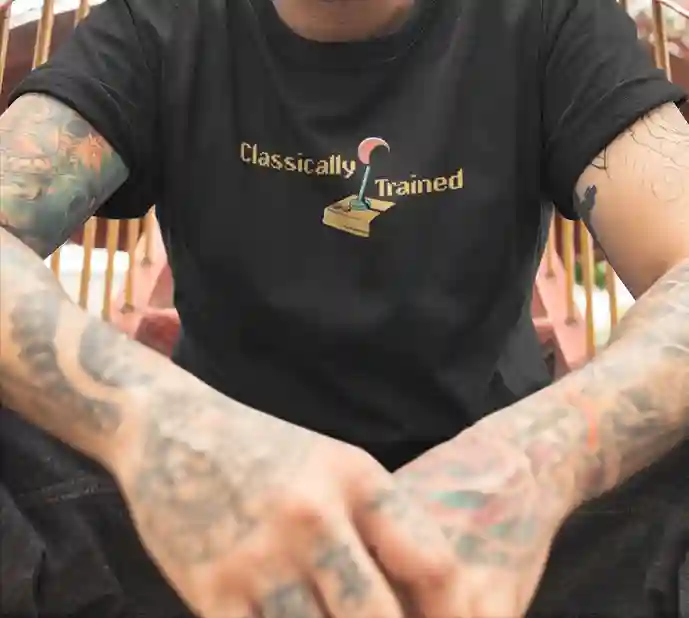 Man wearing a black 'Classically Trained' t-shirt with a retro 80s design featuring an old-school joystick graphic, showing tattooed arms | Satire Clothes