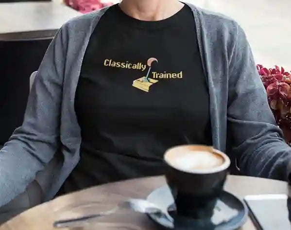 Woman wearing a black 'Classically Trained' t-shirt with a retro 80s design featuring an old-school joystick graphic, sitting at a table with a coffee | Satire Clothes