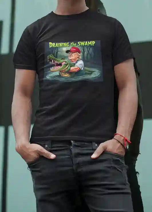 A man wearing a black t-shirt with a graphic design featuring a cartoon Donald Trump in waders holding a large alligator in a swamp under a full moon, with the text 'DRAINING THE SWAMP' above in bold yellow and green letters. | Satire Clothes