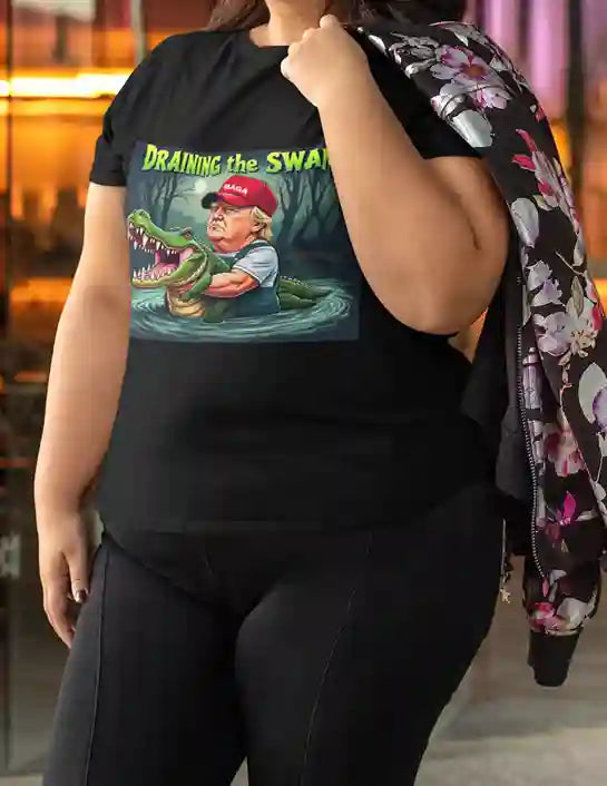 A woman wearing a black t-shirt with a graphic design featuring a cartoon Donald Trump in waders holding a large alligator in a swamp under a full moon, with the text 'DRAINING THE SWAMP' above in bold yellow and green letters. | Satire Clothes