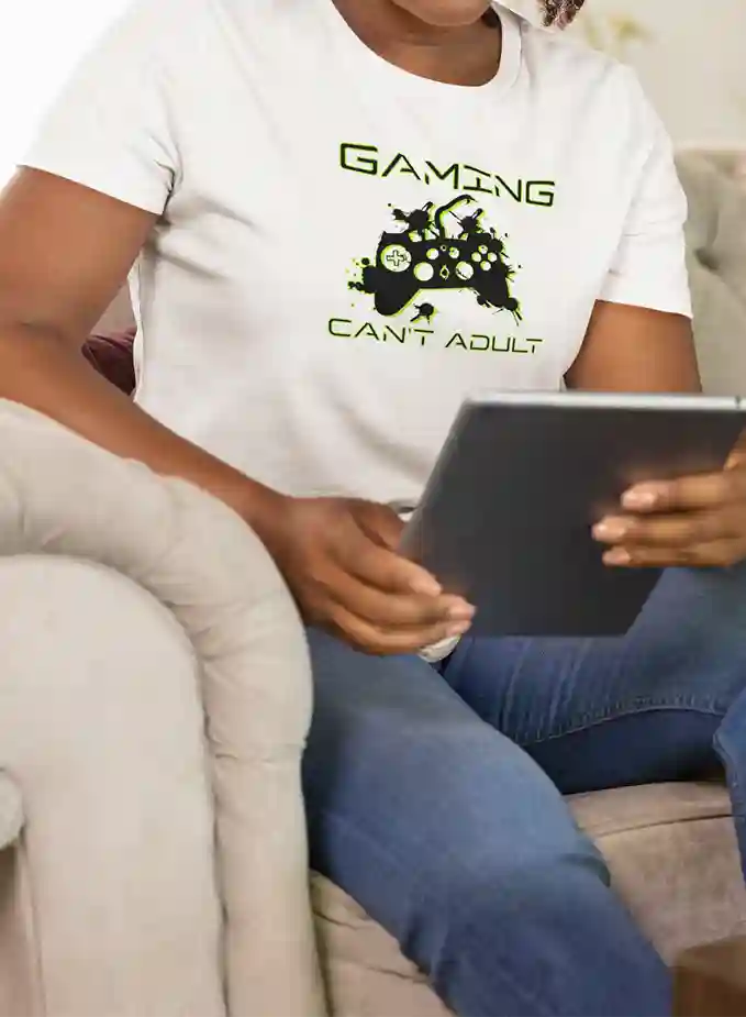 Woman wearing a white T-shirt with the design 'Gaming, Can't Adult' in green and a black silhouette of a gaming controller with green splatter effects | Satire Clothes