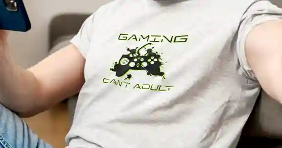 Man wearing a white T-shirt with the design 'Gaming, Can't Adult' in green and a black silhouette of a gaming controller with green splatter effects | Satire Clothes