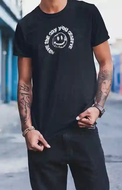 Man wearing a black t-shirt with a distressed smiley face design and the text 'HAVE THE DAY YOU DESERVE' in a circular pattern around it | Satire Clothes