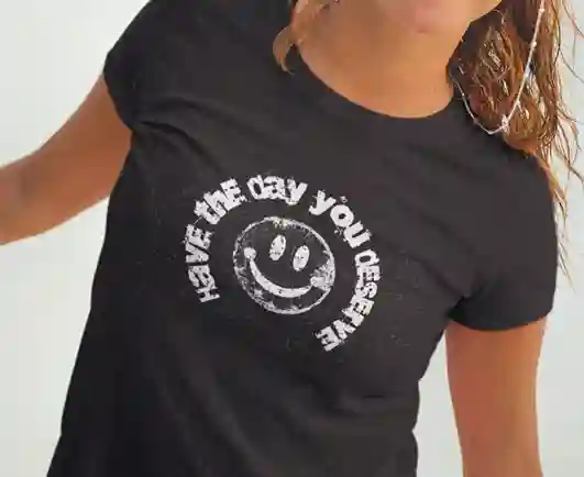 Woman wearing a black t-shirt with a distressed smiley face design and the text 'HAVE THE DAY YOU DESERVE' in a circular pattern around it | Satire Clothes