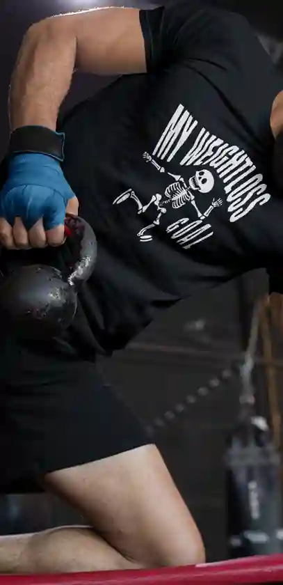 A man wearing a black t-shirt featuring a humorous white skeleton graphic with the text "MY WEIGHTLOSS GOAL" in a distressed font above it | Satire Clothes