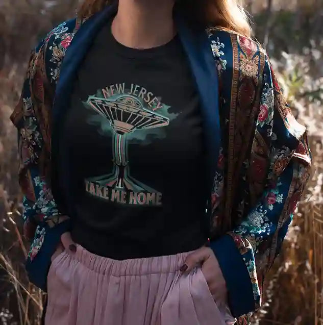 A woman wearing a black t-shirt with a vibrant graphic of a human being abducted by a neon-colored UFO, with the text "NEW JERSEY" above and "TAKE ME HOME" below the image | Satire Clothes