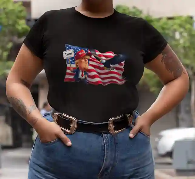 A woman wearing a black t-shirt featuring a satirical design of Donald Trump holding an "I WON" sign in front of an American flag with an eagle flying above | Satire Clothes