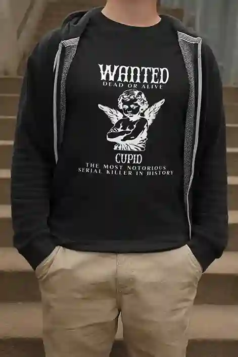 A man wearing a black T-shirt featuring a satirical Wanted: Cupid design, depicting Cupid as the Most Notorious Serial Killer in History | Satire Clothes