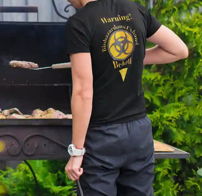 Man wearing a black t-shirt with a humorous warning design on the back. The text reads "Warning! Biohazardous Exhaust" surrounding a biohazard symbol, with "Below" written underneath, all in orange and yellow lettering | Satire Clothes