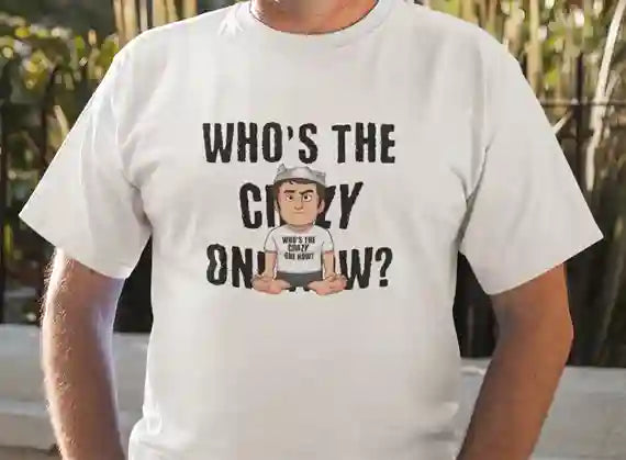 A man wearing a white t-shirt with the text "WHO'S THE CRAZY ONE NOW?" in bold letters. Overlapping the text, there's a cartoon character wearing a tin foil hat and a shirt that says "WHO'S THE CRAZY ONE NOW?" while sitting cross-legged | Satire Clothes