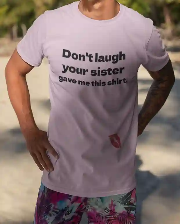 A man wearing a light pink t-shirt with bold black text that reads, "Don't laugh your sister gave me this shirt." At the bottom, there's a red lipstick kiss print. | Satire Clothes